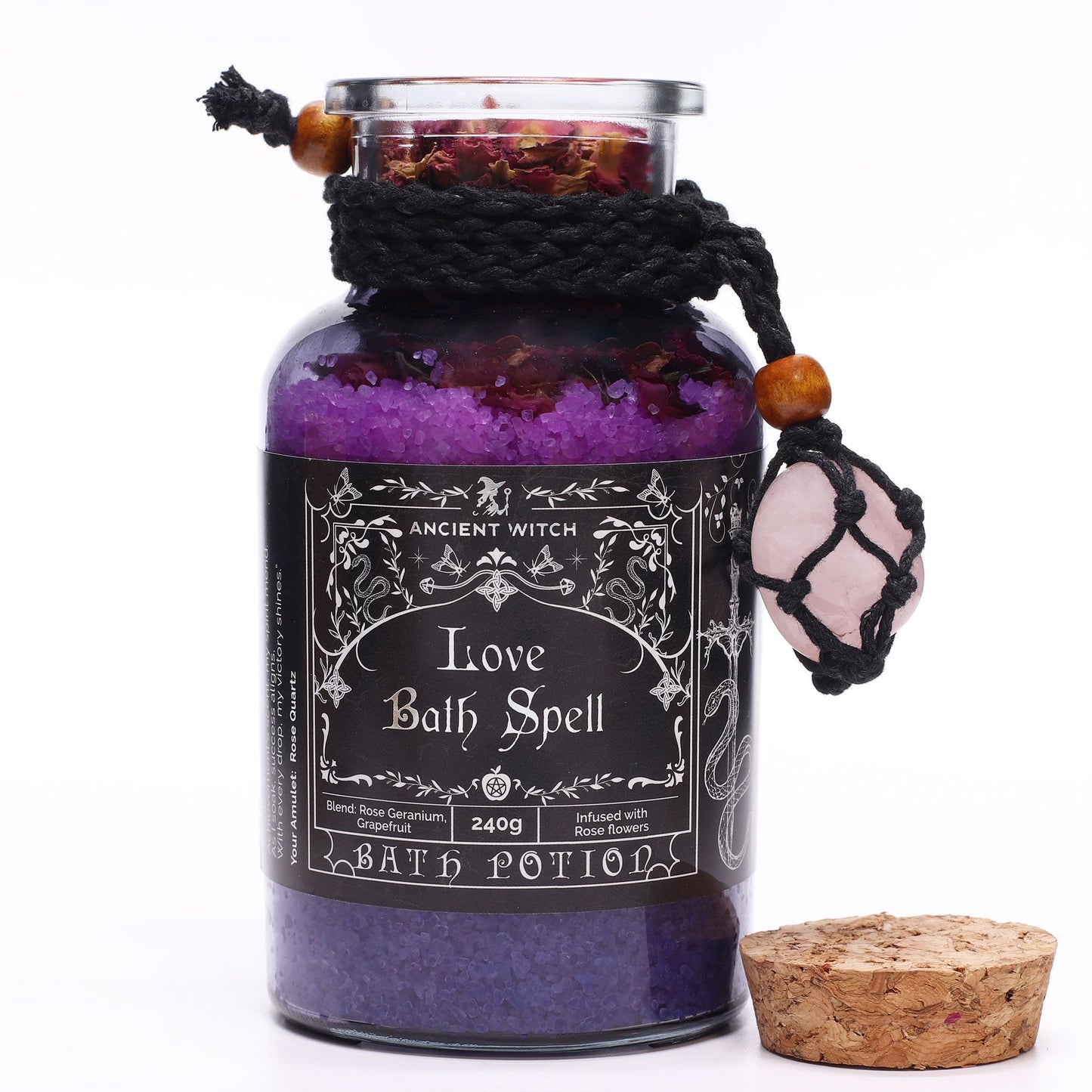 Glass bottle with cork lid off filled with Himalayan bath salts and rose petals infused with Rose Geranium and Grapefruit essential oils with a Rose Quartz crystal amulet
